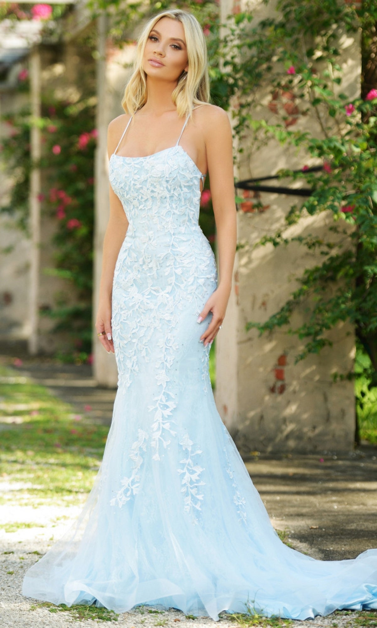 A woman stands outdoors in the Beaded Lace Long Prom Dress Dutchess, a light blue, floor-length gown with thin straps. Pink flowers and greenery in the background enhance the elegance of this designer formal dress.